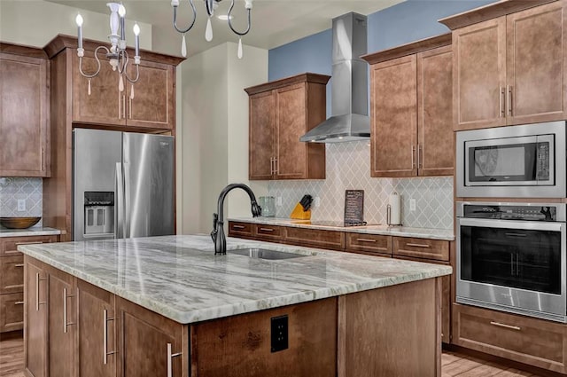 kitchen with light stone countertops, a kitchen island with sink, a sink, stainless steel appliances, and wall chimney range hood