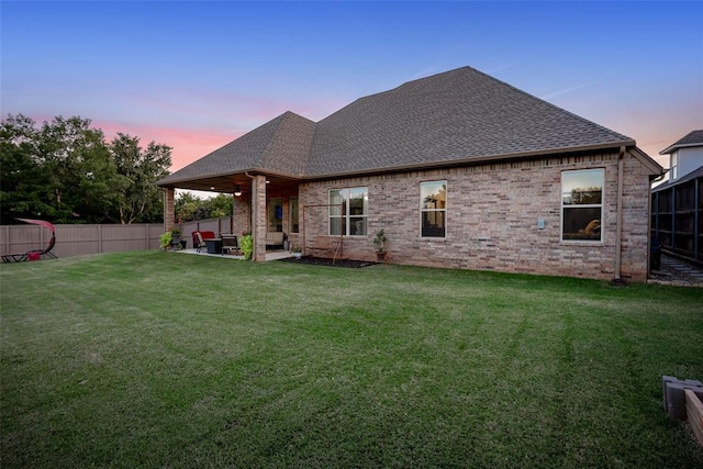 back of property at dusk with brick siding, roof with shingles, a lawn, a fenced backyard, and a patio area