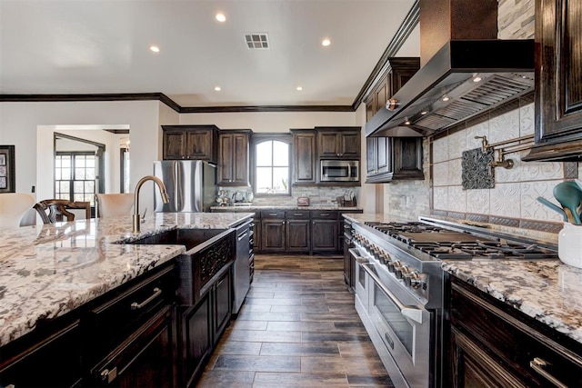 kitchen featuring stainless steel appliances, wall chimney exhaust hood, a sink, dark brown cabinets, and light stone counters