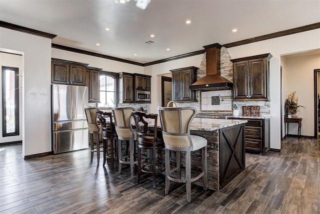 kitchen with appliances with stainless steel finishes, wall chimney range hood, dark brown cabinets, a large island, and ornamental molding