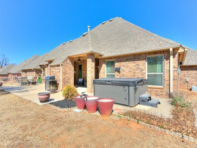 rear view of house with a shingled roof, a patio area, brick siding, and a hot tub