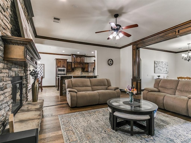 living room with visible vents, ceiling fan with notable chandelier, wood finished floors, a stone fireplace, and crown molding