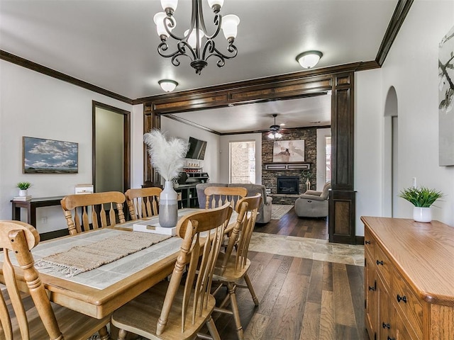 dining space with arched walkways, dark wood-style flooring, ornamental molding, a stone fireplace, and ceiling fan with notable chandelier