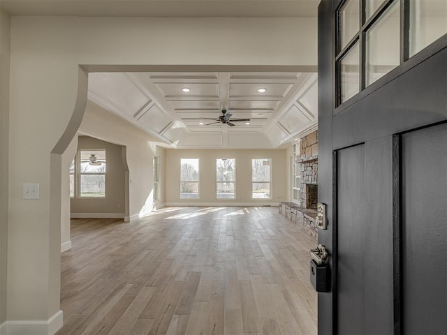 unfurnished living room with light wood-type flooring, a ceiling fan, coffered ceiling, a stone fireplace, and baseboards