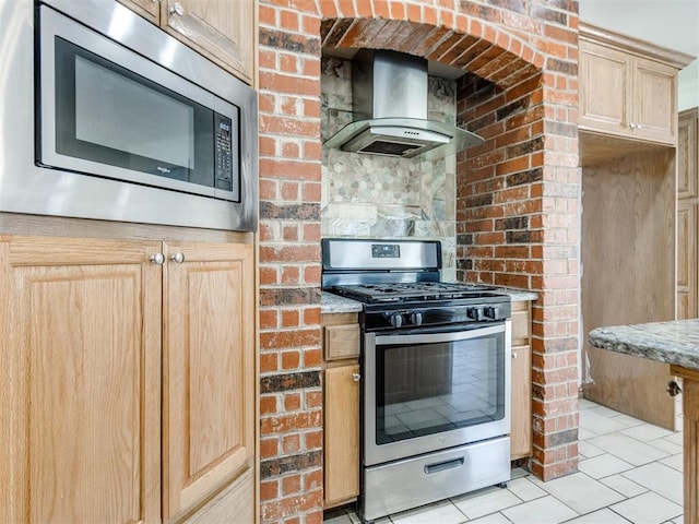 kitchen featuring brick wall, light tile patterned flooring, light brown cabinetry, appliances with stainless steel finishes, and wall chimney range hood