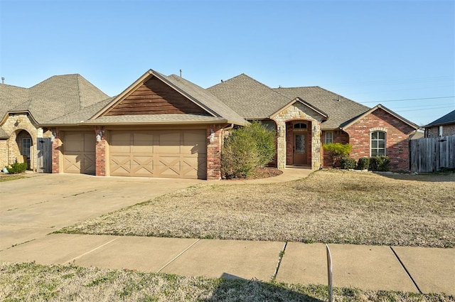 french country inspired facade with brick siding, fence, roof with shingles, a garage, and driveway