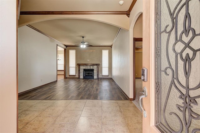 unfurnished living room with a ceiling fan, arched walkways, a stone fireplace, crown molding, and tile patterned flooring