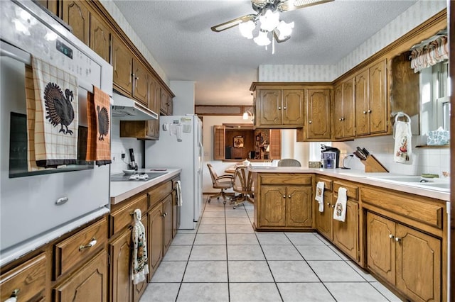 kitchen featuring ceiling fan, under cabinet range hood, light countertops, brown cabinets, and a peninsula