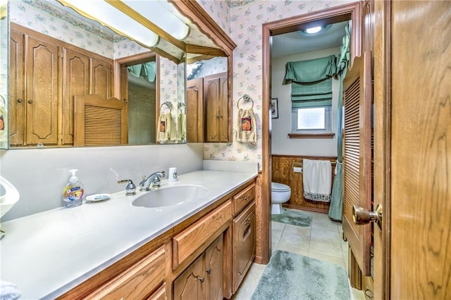 bathroom featuring tile patterned floors, a wainscoted wall, toilet, wallpapered walls, and vanity