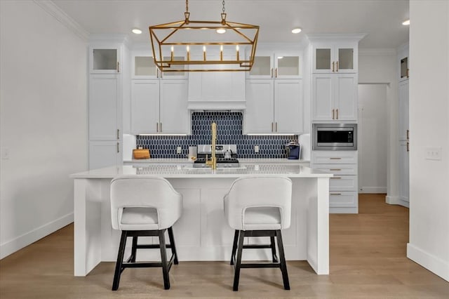 kitchen featuring stainless steel microwave, an island with sink, light countertops, ornamental molding, and decorative backsplash