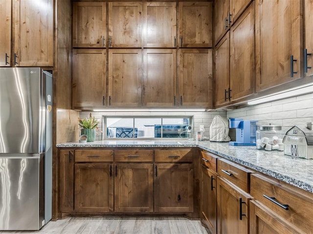 kitchen featuring brown cabinetry, light stone countertops, light wood-type flooring, freestanding refrigerator, and tasteful backsplash