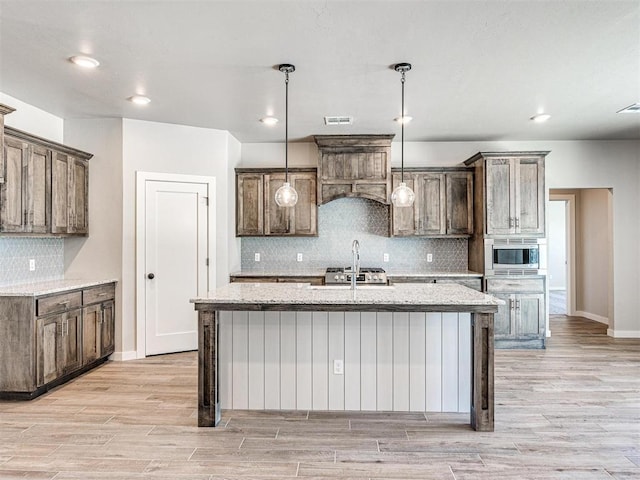 kitchen with an island with sink, light wood-style flooring, a sink, stainless steel microwave, and decorative light fixtures