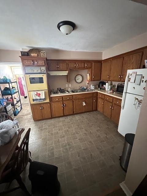 kitchen with under cabinet range hood, light countertops, brown cabinetry, white appliances, and a sink