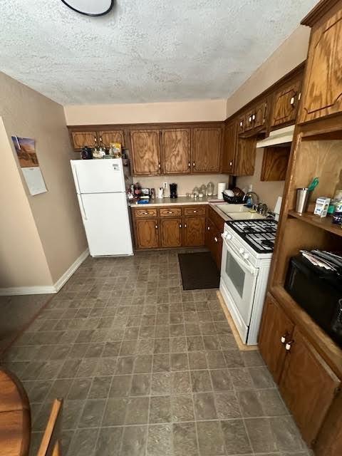 kitchen featuring white appliances, under cabinet range hood, a textured ceiling, a sink, and baseboards
