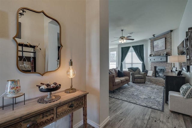 living room featuring wood finished floors, a ceiling fan, visible vents, baseboards, and a brick fireplace