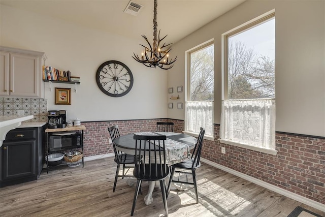 dining space with a wainscoted wall, visible vents, brick wall, light wood-style floors, and a notable chandelier