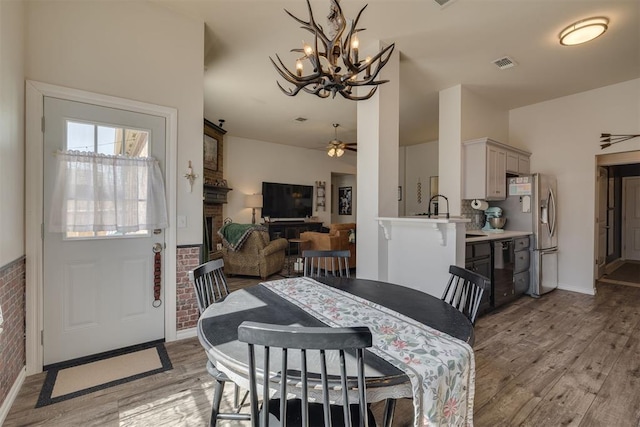 dining space featuring light wood-type flooring, visible vents, brick wall, and ceiling fan with notable chandelier