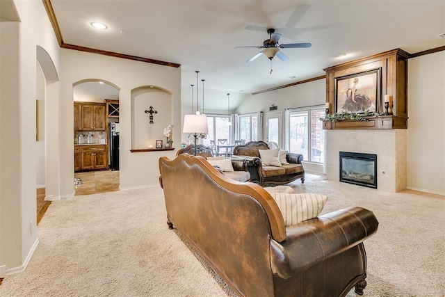 living area featuring ornamental molding, a ceiling fan, a large fireplace, arched walkways, and light colored carpet