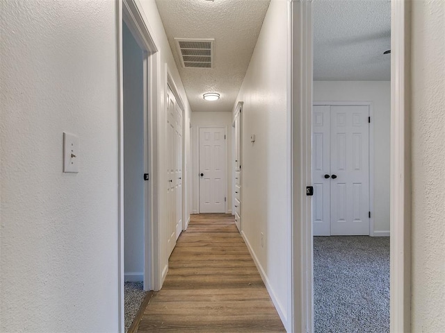 hall featuring wood finished floors, baseboards, visible vents, a textured ceiling, and a textured wall