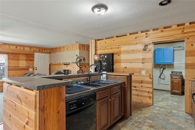 kitchen featuring wooden walls, a sink, stone finish floor, black appliances, and a kitchen island with sink