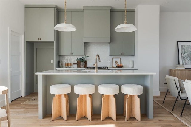 kitchen featuring light wood-type flooring, an island with sink, a sink, backsplash, and light countertops