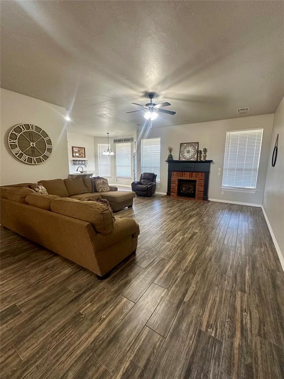 unfurnished living room featuring a textured ceiling, dark wood finished floors, baseboards, a brick fireplace, and ceiling fan