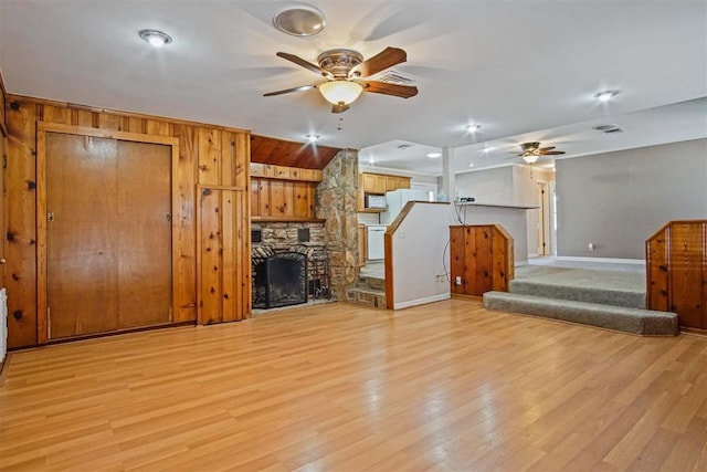 unfurnished living room with stairway, a stone fireplace, ceiling fan, and light wood-style flooring