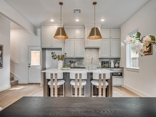 kitchen with light wood finished floors, visible vents, a sink, backsplash, and a center island with sink