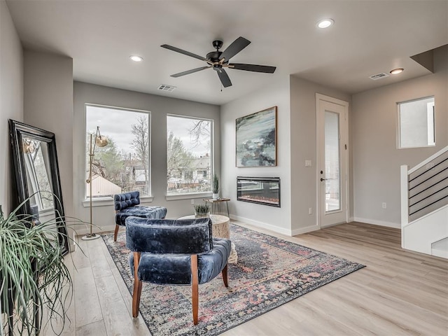 living area with stairs, visible vents, a ceiling fan, a glass covered fireplace, and wood finished floors