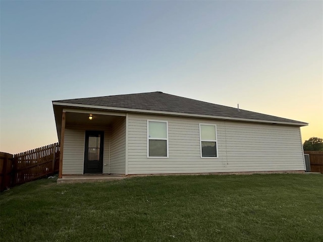 back of property at dusk with a lawn, fence, and roof with shingles