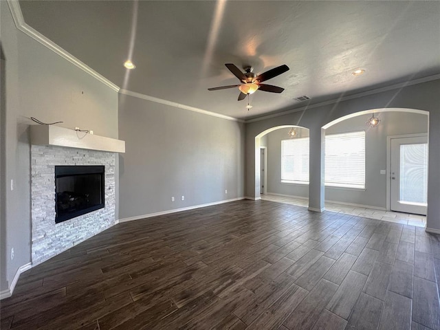 unfurnished living room featuring a ceiling fan, dark wood-style flooring, crown molding, a stone fireplace, and arched walkways