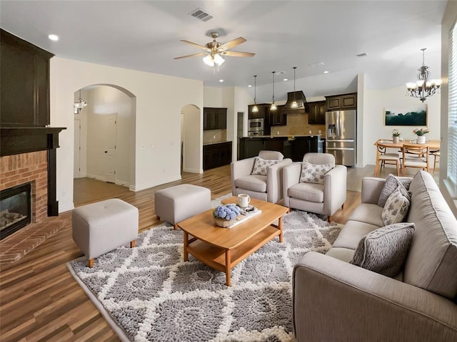 living room featuring visible vents, a brick fireplace, light wood finished floors, arched walkways, and ceiling fan with notable chandelier