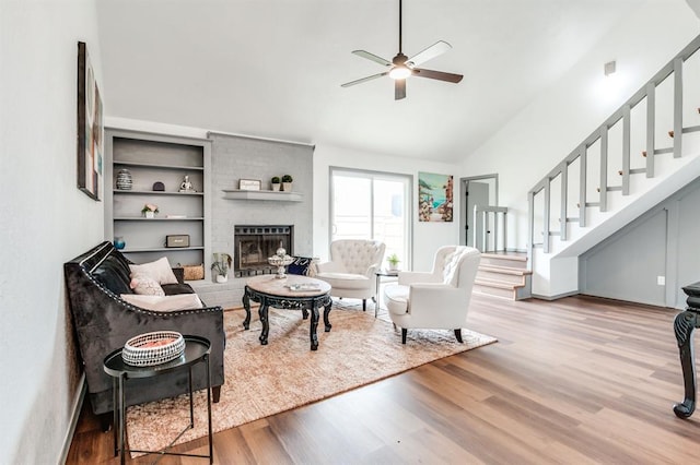 living room with high vaulted ceiling, wood finished floors, stairs, ceiling fan, and a brick fireplace