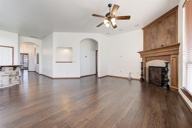 unfurnished living room featuring dark wood-type flooring, baseboards, a large fireplace, arched walkways, and ceiling fan