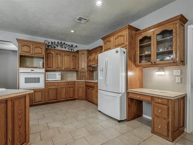 kitchen featuring visible vents, built in desk, white appliances, brown cabinetry, and light countertops