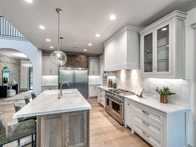 kitchen featuring high end appliances, a sink, arched walkways, wall chimney exhaust hood, and glass insert cabinets