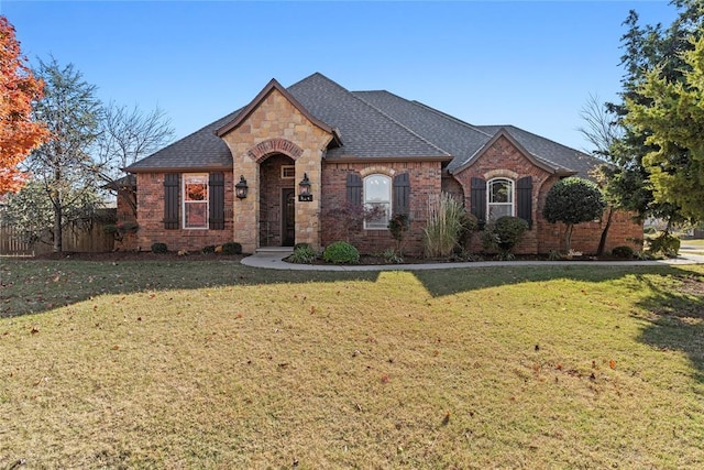 french country home featuring stone siding, fence, a front yard, brick siding, and a shingled roof
