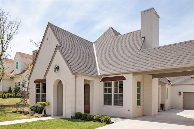 tudor home featuring roof with shingles, a chimney, concrete driveway, brick siding, and a garage
