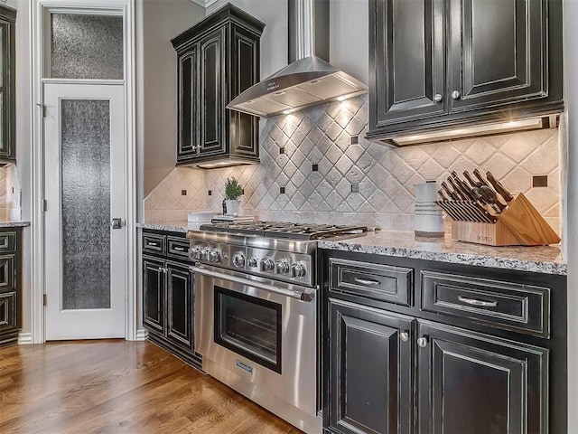 1109 Salvo Bridge Ct, Edmond, OK 73034 - kitchen featuring wall chimney exhaust hood, high end stainless steel range, dark cabinets, light wood finished floors, and light stone countertops