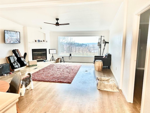 living room with ornamental molding, wood finished floors, a glass covered fireplace, and a ceiling fan