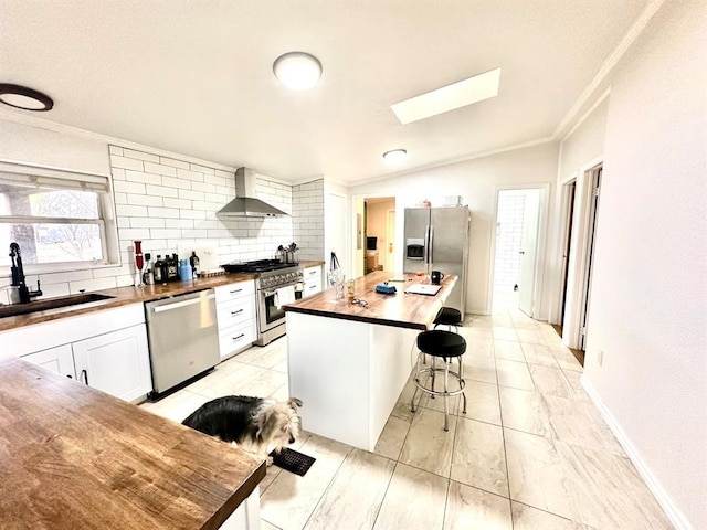 kitchen with a sink, wall chimney exhaust hood, a kitchen breakfast bar, appliances with stainless steel finishes, and butcher block counters