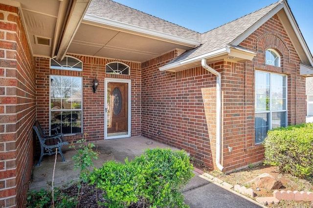 view of exterior entry with brick siding, visible vents, and roof with shingles