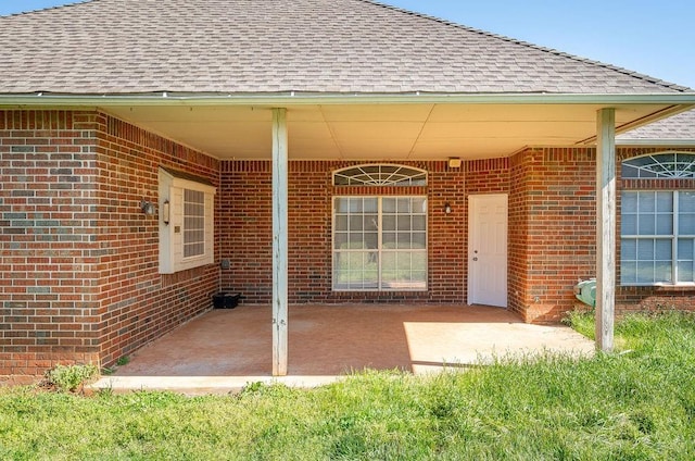view of exterior entry with a shingled roof, a patio area, and brick siding