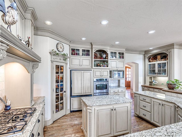 kitchen featuring ornamental molding, stainless steel appliances, arched walkways, light wood finished floors, and glass insert cabinets