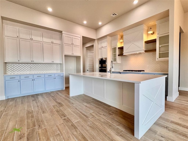 10132 NW 100th St, Yukon, OK 73099 - kitchen featuring open shelves, white cabinets, an island with sink, light wood-style flooring, and light countertops