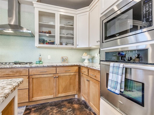 kitchen featuring tasteful backsplash, glass insert cabinets, wall chimney range hood, appliances with stainless steel finishes, and light stone counters