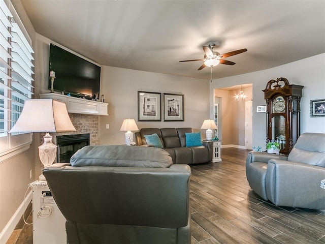 living room featuring ceiling fan with notable chandelier, baseboards, a fireplace, and dark wood-style flooring