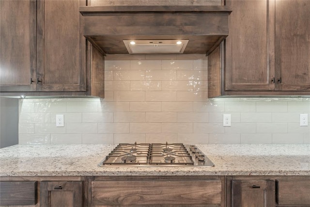 kitchen with light stone countertops, tasteful backsplash, dark brown cabinets, stainless steel gas cooktop, and custom exhaust hood