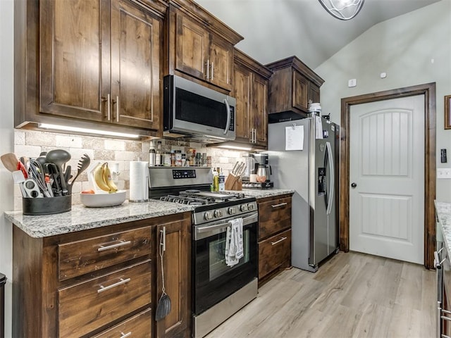 kitchen with light wood-type flooring, vaulted ceiling, light stone countertops, decorative backsplash, and appliances with stainless steel finishes