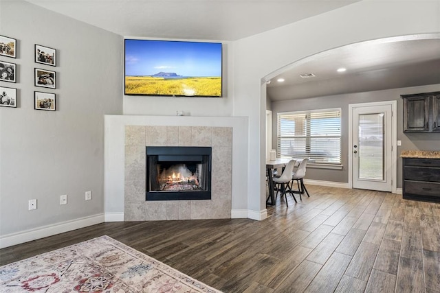 living room featuring arched walkways, visible vents, wood finished floors, a tiled fireplace, and baseboards